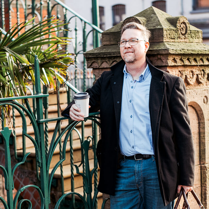 JD Gibson voiceover stood in front of iron railings holding a mug of coffee.